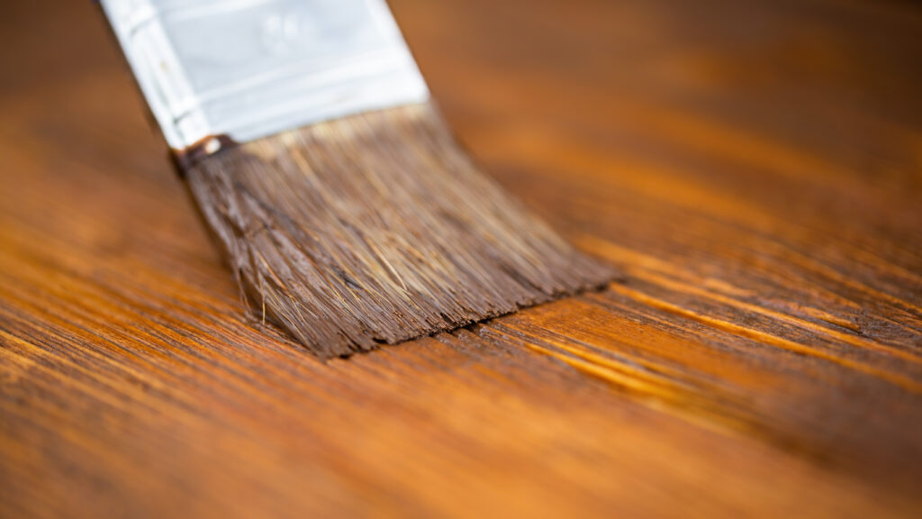 Close-up of paintbrush applying brown color on wooden table. Detail of working tool painting a color on surface. Repairing dark desk with protection oil.